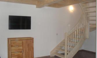 A bright room with wooden flooring and a curved staircase. A television hangs on the wall above a wooden cabinet.