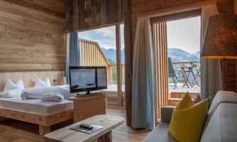 A cozy room with wooden furniture and large windows. On the balcony is a terrace with a view of the mountains and a small table.