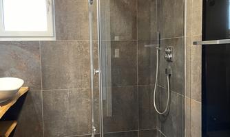 A modern shower cubicle with glass walls and an elegant design. The room has gray tiles and a large window.
