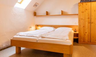 A cozy bedroom with a wooden bed and two mattresses. There is a wooden wardrobe and a window that lets in natural light.