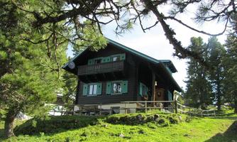 An old, charming wooden house in the mountains, surrounded by green meadows and trees. The sky is clear and sunny.