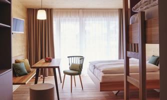 A modern room with wooden decor, a double bed, and a bunk bed. At a table, there are two chairs and a small stool.