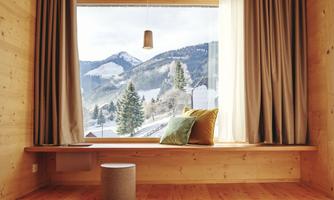 A bright wooden room with a large window that offers a view of snowy mountains and trees. The room features cozy cushions and a small stool.