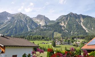 A picturesque mountain landscape with high peaks and lush greenery. In the foreground, blooming plants and a peaceful residential area.