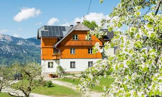 A picturesque house with orange and white walls, surrounded by blossoming trees. In the background, the mountains and a clear sky can be seen.