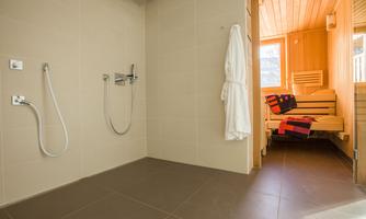 A modern bathroom with a shower and a white bathrobe. In the background, a sauna with a wooden bench is visible.