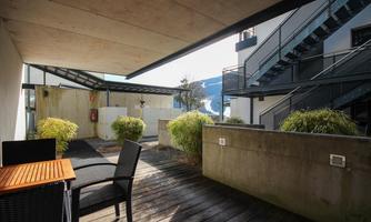 A modern outdoor area with a wooden terrace and a table chair. In the background, there are plants and a metal staircase visible.