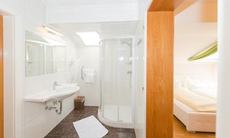 A modern bathroom with a shower, toilet, and sink. Next to the bathroom there is a bedroom with a large bed.