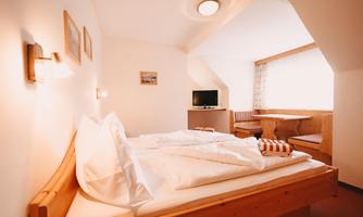 A cozy room with a double bed and warm wooden paneling. There is a table and a television, as well as a large window that provides natural light.