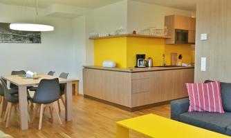 A modern apartment with a bright kitchen and a dining area. The living area is cozy with a sofa and a colorful table.