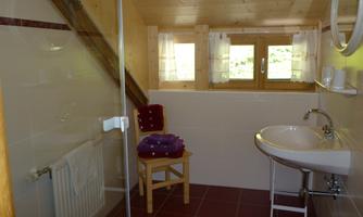 A modern bathroom with a glass shower and a sink. There are large windows that let in a lot of light, and towels on a wooden chair.
