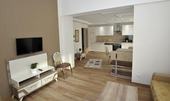 A modern living room with wooden flooring and a television. In the background, there is an open kitchen with white cabinets.