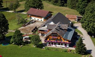 A picturesque farmhouse surrounded by green meadows and trees. The buildings have charming balconies with flowers, and there is a peaceful rural setting.