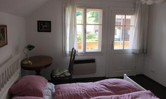 A cozy bedroom with a double bed and checkered blankets. There is a table, a chair, and large windows overlooking nature.