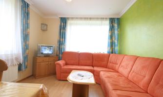 A cozy living room with an orange couch and a table. The walls are painted green and large windows let in plenty of light.