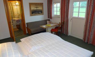 A cozy room with two beds, a small table, and a seating area. In the background, the bathroom and a window with curtains can be seen.