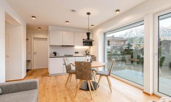 A modern kitchen with a dining area and large windows. The room has hardwood flooring and offers a view of a green landscape.