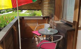 A cozy balcony with a colorful sun umbrella, a small table, and two chairs. The surroundings are green and inviting.