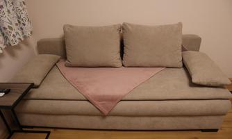 A beige sofa with two large cushions and a pink blanket. Next to the sofa is a small table.