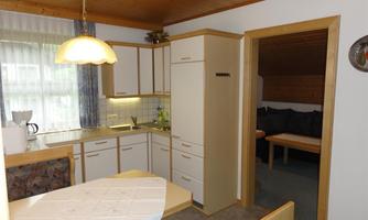A modern kitchen with a table and chairs. In the background, a living room with a couch is visible.