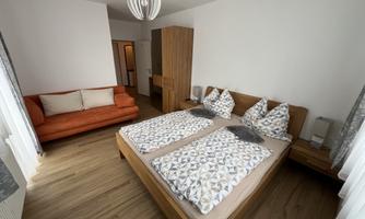 A cozy bedroom with a large bed and colorful pillows. The room features an orange sofa and light wood design.