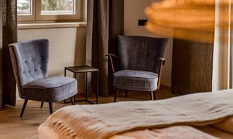 A cozy room with two blue armchairs and a small table. Soft light falls through the window onto the wooden floor and the warm textiles.