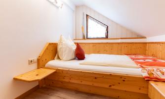 A cozy bedroom with a wooden bed and two pillows. The walls are light, and there is a small window with a view outside.