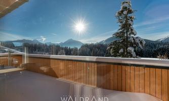 A beautiful winter landscape with snow-covered mountains and bright sunshine. The view from a balcony shows a calm, picturesque surroundings.