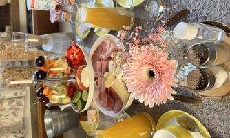 A hearty breakfast with fresh fruit, cold cuts, vegetables, and juice. The table is beautifully set with dishes and a blooming decoration.