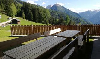 A beautiful terrace with wooden tables and benches overlooks an impressive mountain landscape. In the background, green forests and snow-capped peaks can be seen.