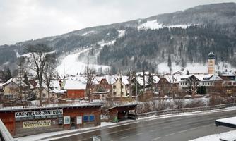 A snowy landscape with a small village and mountains in the background. The streets are empty and the buildings are covered with snow.