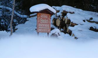 A wooden sign with the inscription "Guesthouse Sprott" stands in the snow. In the background, stacked logs can be seen.