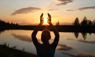 A person stands on the shore of a calm lake, holding their hands above their head to embrace the sun. The sunset colors the sky in warm hues.