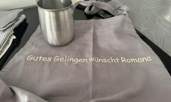 A gray chef's apron with the inscription "Wishing you good success, Romana" lies on a work surface. Next to it stands a stainless steel measuring cup.