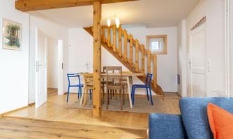 A cozy living space with a wooden staircase and a dining table. The decor is modern and inviting with colorful chairs.