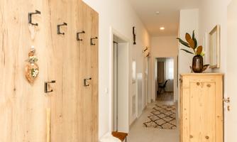 A modern hallway with wooden panels and several coat hooks. On the right side stands a beautiful cabinet, and the floor is tiled with light tiles.
