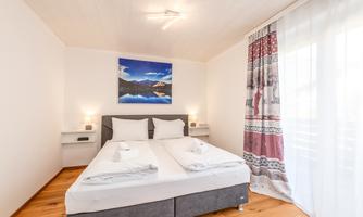 A cozy bedroom with a double bed and modern furniture. A landscape painting hangs on the wall, and a bright window provides daylight.