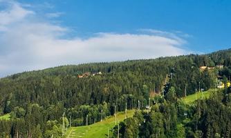 A green mountain landscape with trees and a clear blue sky. Some cable car supports are visible.