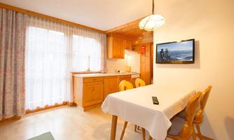 A bright kitchen with a wooden dining area and a television on the wall. Large windows provide plenty of daylight.
