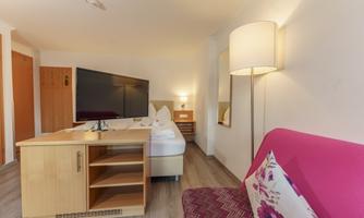 A bright hotel room with a large TV, a single bed, and a cozy sofa. The decor is simple and modern, with warm light sources.