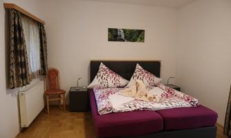 A cozy bedroom with a double bed and colorful pillows. A chair and a window with curtains complement the space.