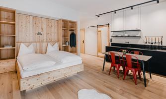 A modern room with a wooden double bed and a cozy dining area. The kitchen is dark-colored and well-equipped.