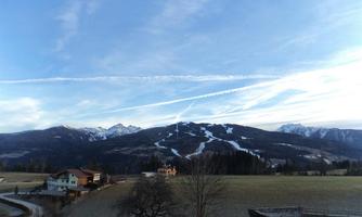 A picturesque landscape with snow-covered mountains and a clear sky. In the foreground, green meadows and a small village can be seen.