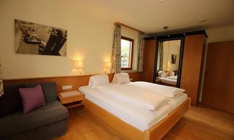 A cozy bedroom with a double bed and a gray sofa. Large windows provide bright light and a relaxed atmosphere.