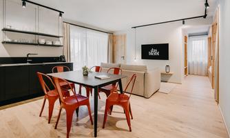 A modern kitchen-living room with a table and red chairs. The space is bright and welcoming, with large windows and a cozy couch.