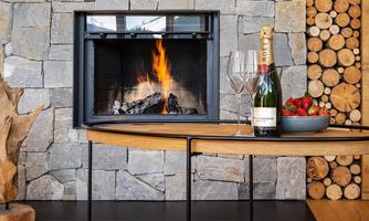 A cozy fireplace with a flaming fire in front of a stone wall. On the table are champagne, glasses, and a bowl of strawberries.