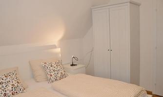 A cozy bedroom with a double bed and colorful cushions. The room is decorated in light tones and has a white wardrobe.