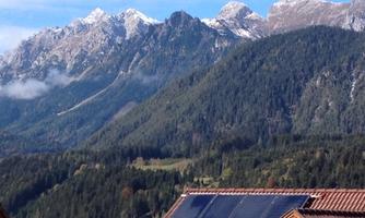 A beautiful mountain panorama with snow-capped peaks and dense forests. In the foreground, solar panels are visible on a roof.