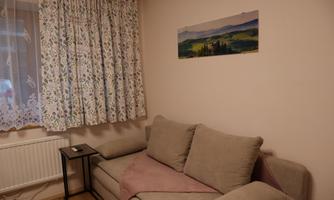 A cozy living room with a beige sofa and a small table. The windows are adorned with a floral patterned curtain.
