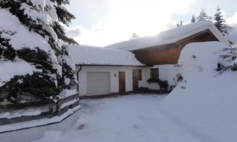 A wintery house, surrounded by snow and fir trees. The sky is bright and cloudy.